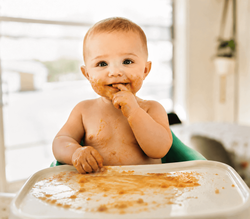 Baby isst Beikost im Hochstuhl und ist mit Essen bekleckert